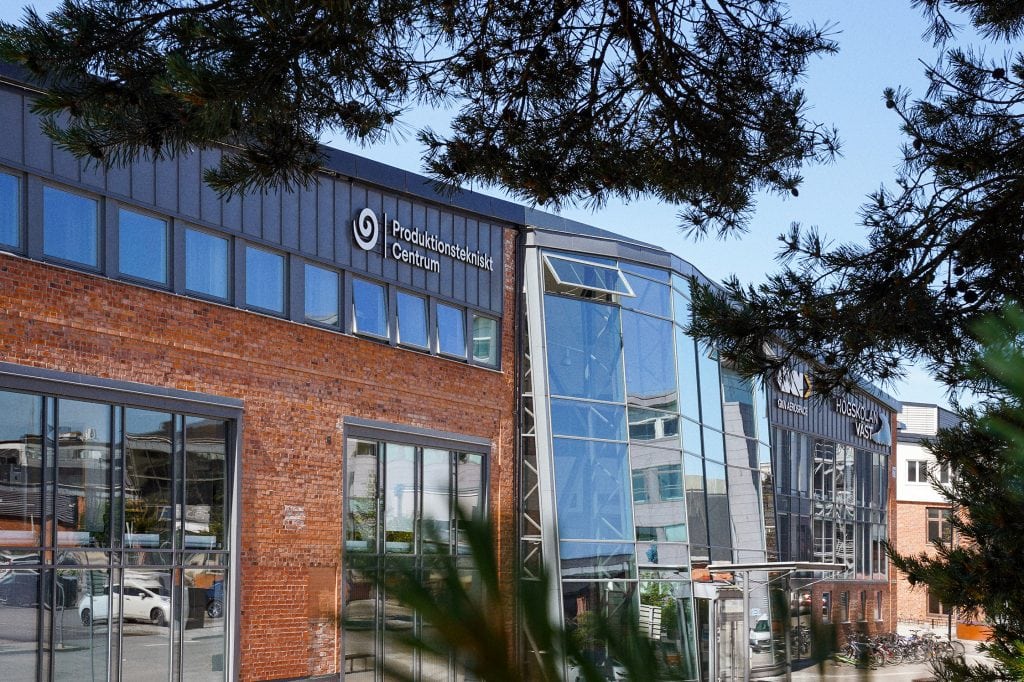 Utsidan av PTC fasad. Tallgrenar i förgrunden. Tegelbyggnad och stora glaspartier.