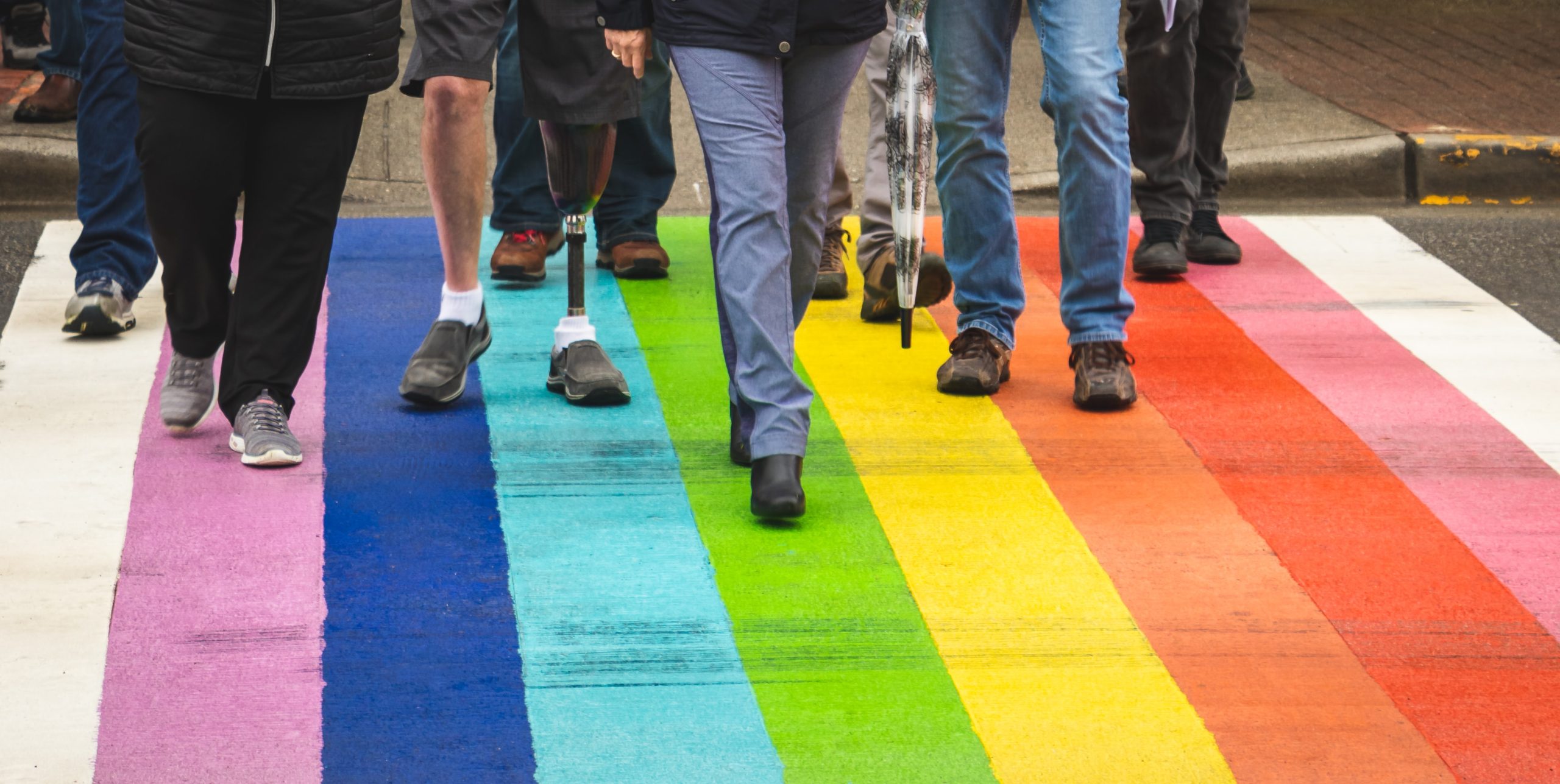 Olika människor går på övergångsställe målat i regnbågsfärger. Bara nederdelen av kropparna syns. En person går med kryckor, en annan person har en benprotes.